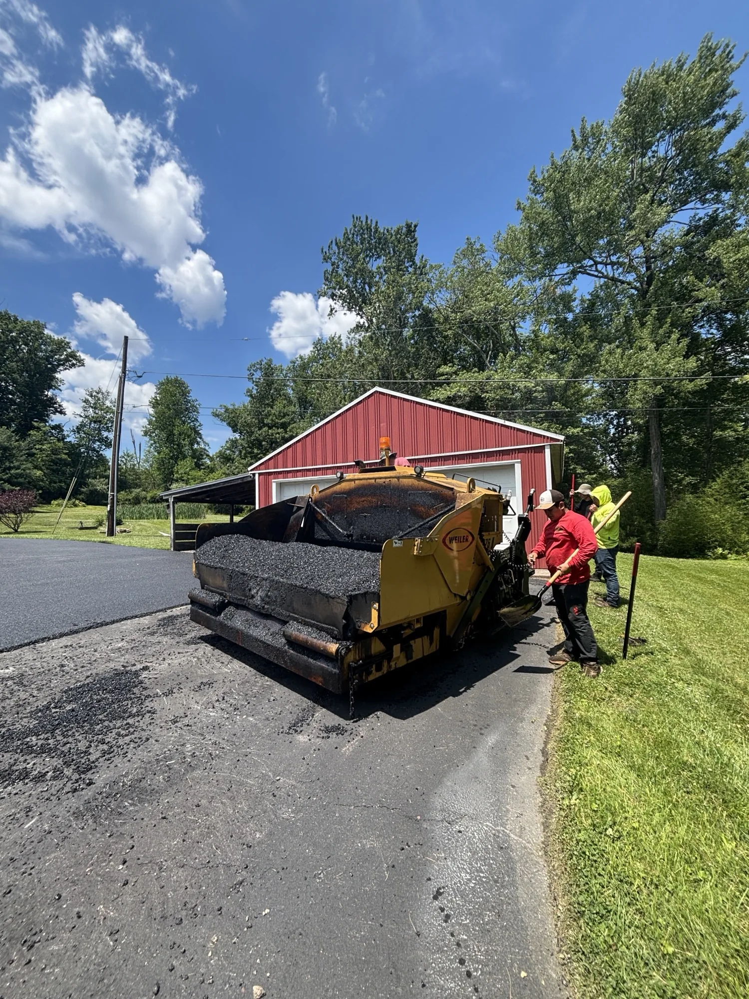 Yellow paver and All County crew dropping a fresh load of hot-mix asphalt near a red barn property